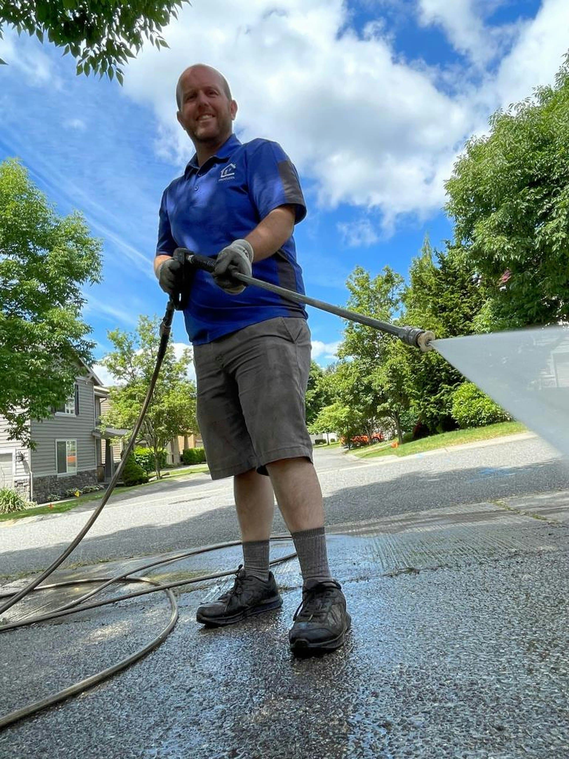 Man in a blue shirt pressure washing a concrete driveway in front of a house